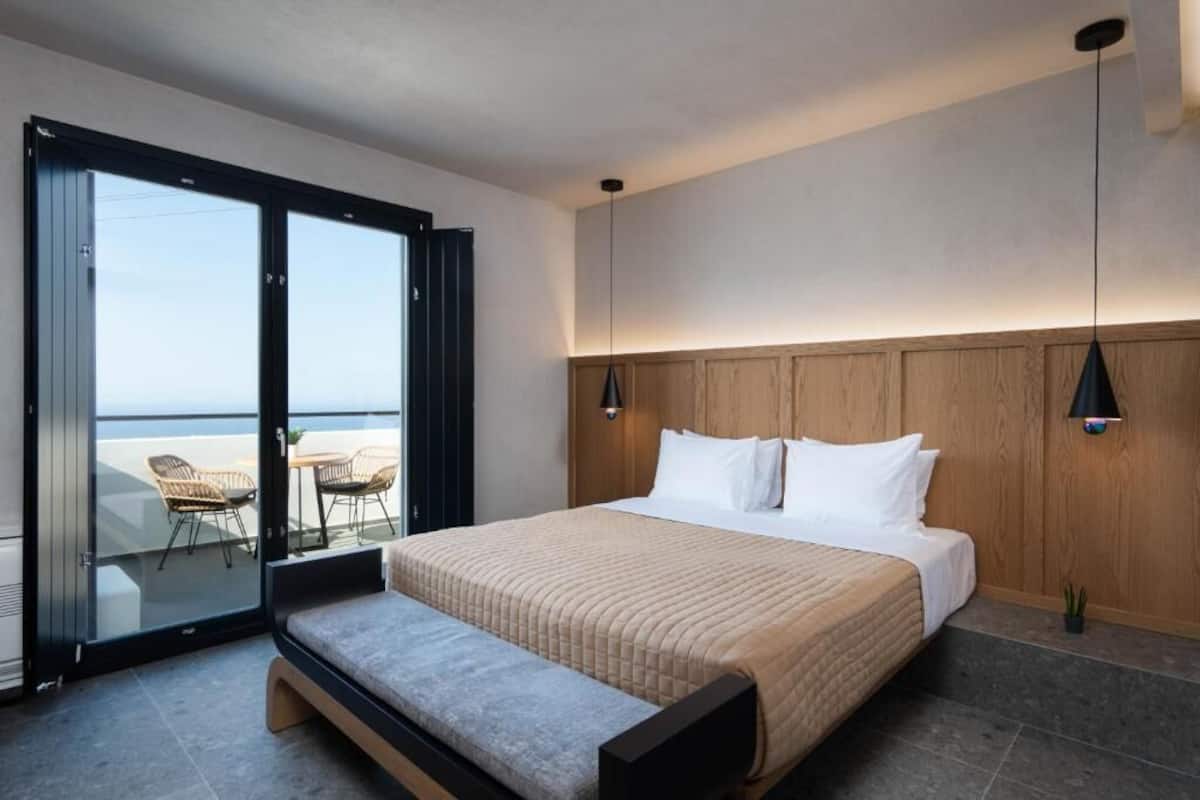 Ein modernes Schlafzimmer mit einem großen Bett, einer Sitzbank und einer Schiebetür, die auf einen Balkon mit Stühlen führt.