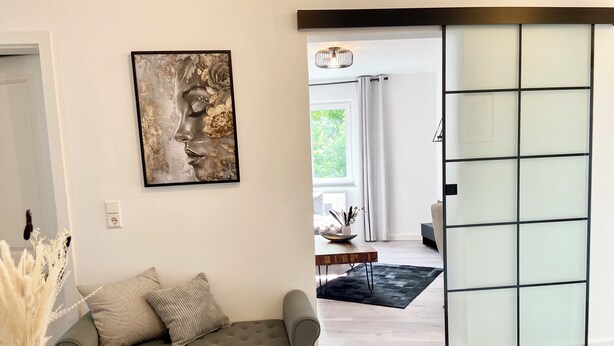 Ein modernes Wohnzimmer mit einem grauen Sofa, einem gerahmten abstrakten Gemälde und einer Glaswand, die in einen anderen Raum führt.