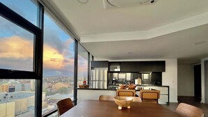 Un comedor moderno con una mesa grande y sillas, una cocina al fondo y vistas a la ciudad a través de amplias ventanas.