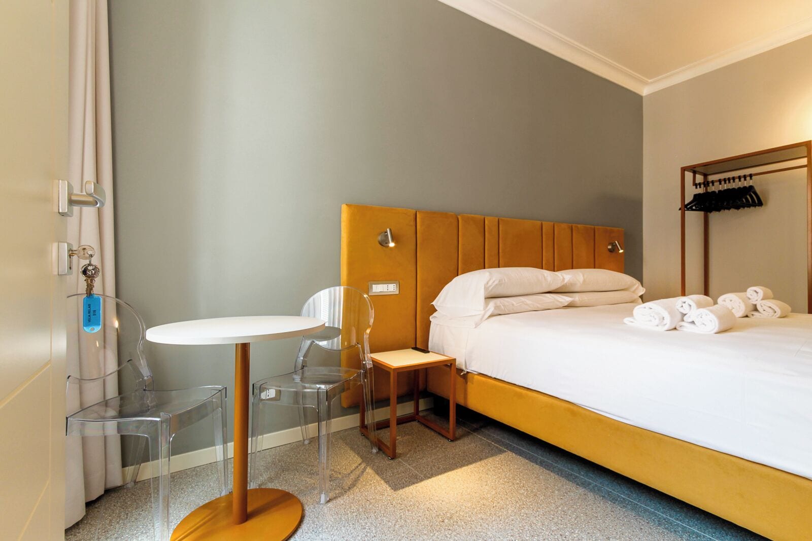 A hotel room with a bed, a small table, two chairs, a coat rack, and a bathroom door.