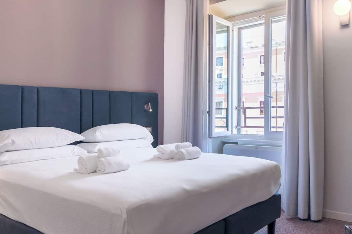A neatly made bed with white linens and towels, a dark blue headboard, and a window with curtains.