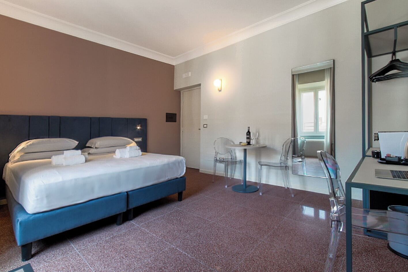 Habitación de hotel con cama, escritorio con computadora, silla y pequeña mesa con botella y vasos.