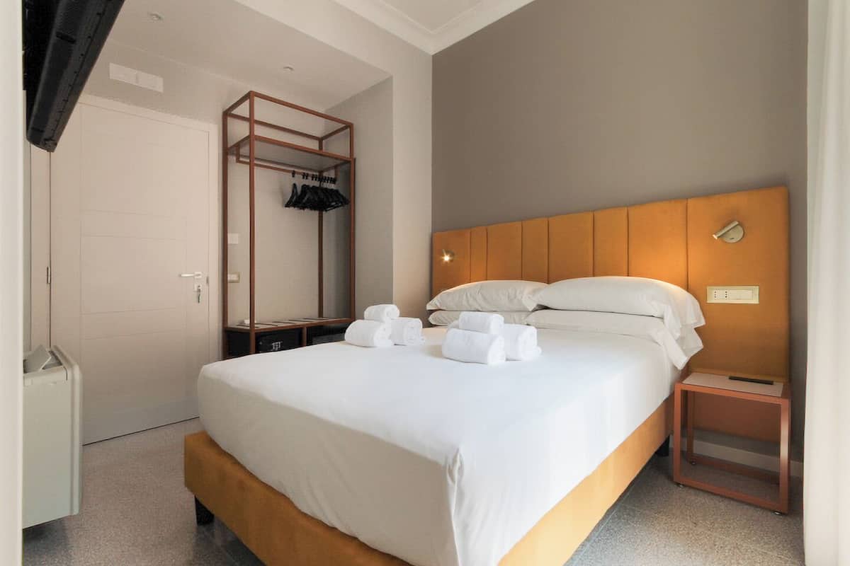 A hotel room with a bed, a wooden headboard, a bedside table, a nightstand, a wardrobe, and a mirror.