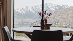 Een kamer met een groot raam met uitzicht op een bergachtig landschap. Er staat een tafel met een vaas en een schaal erop, en er zijn twee stoelen voor de tafel.