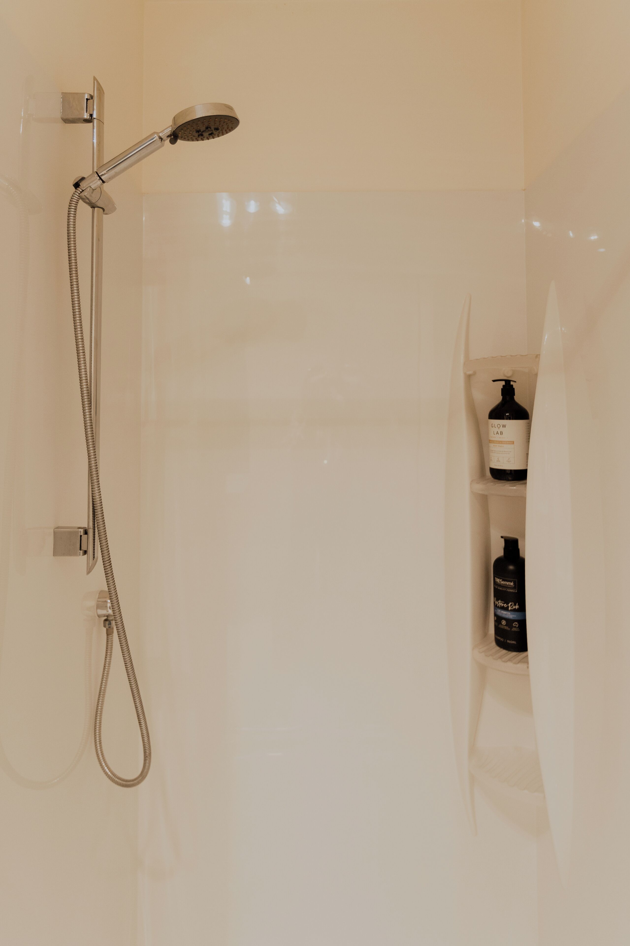 A shower with a handheld showerhead and a flexible hose. There are two bottles of shampoo and conditioner on a shelf.