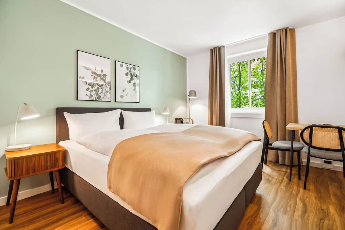 Een hotelkamer met een groot bed, twee nachtkastjes, een bureau met stoel, en een raam met gordijnen.