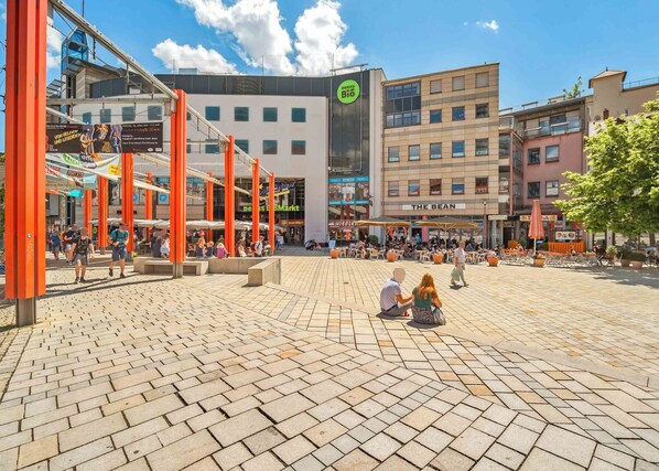 An outdoor public square with a modern building featuring a green logo, surrounded by cafes and shops. - Limehome Jena Teichgraben (Jena)