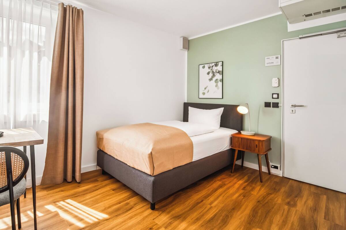 Een netjes ingerichte hotelkamer met een bed, nachtkastje, lamp en uitzicht buiten door dunne gordijnen.