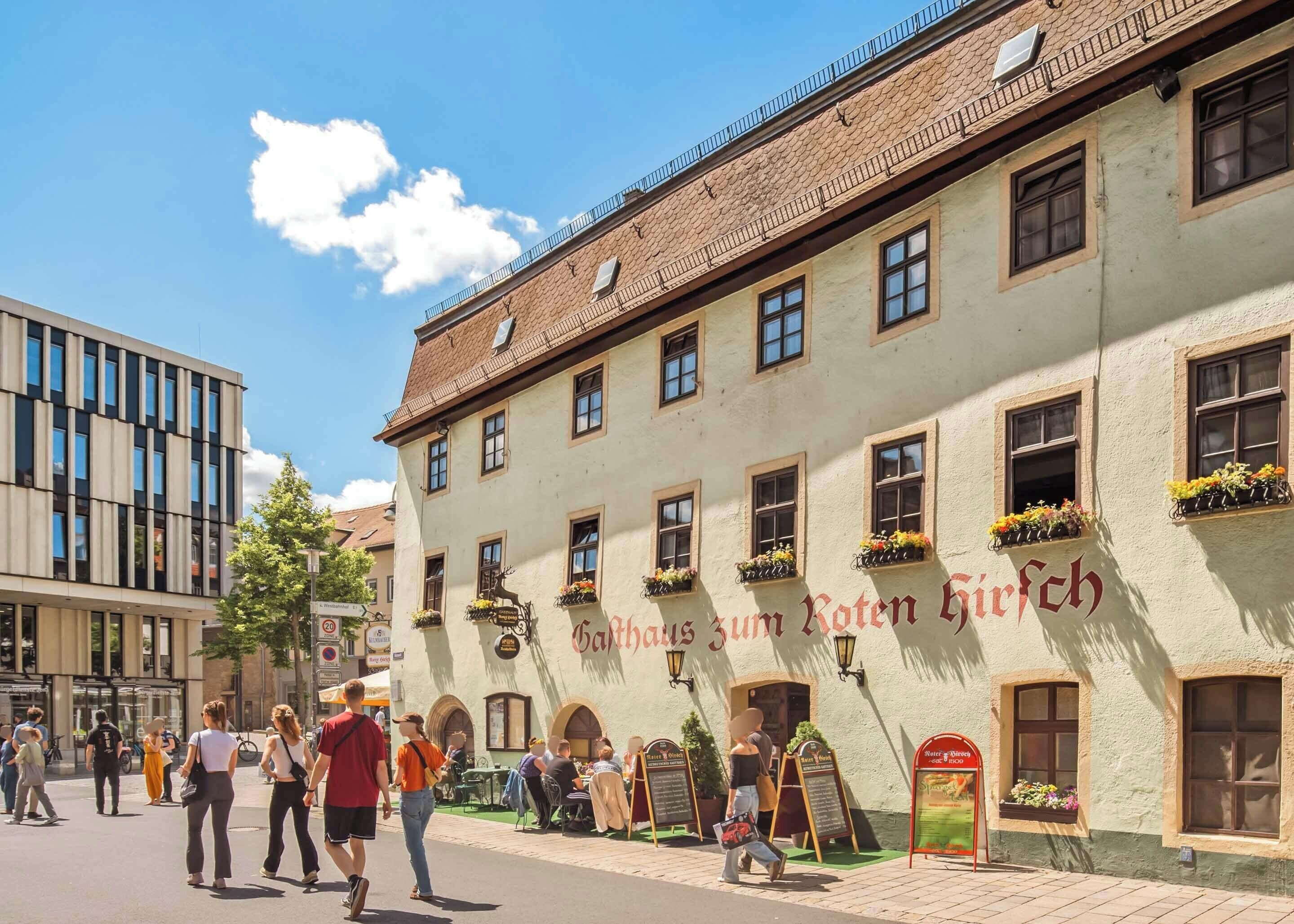 A traditional building with a sign reading "Gasthof zum Roten Hirsch" and outdoor seating with flower baskets.