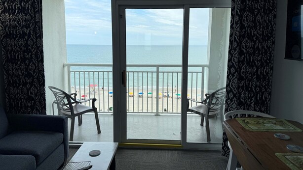 A room with a balcony, a sofa, a table, and a view of the beach and ocean. - Amazing views - Kings size Ocean front Camelot 715 (Myrtle Beach)