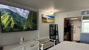 A hotel room with a bed, a dresser, a TV displaying a mountain landscape, a painting of a beach, and a kitchenette area. - Amazing views - Kings size Ocean front Camelot 715 (Myrtle Beach)