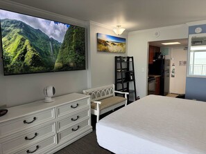 A hotel room with a bed, a dresser, a TV displaying a mountain landscape, a painting of a beach, and a kitchenette area. - Amazing views - Kings size Ocean front Camelot 715 (Myrtle Beach)