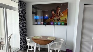 A hotel room with a TV mounted on the wall displaying various streaming services, a wooden table with chairs, and a window with a view. - Amazing views - Kings size Ocean front Camelot 715 (Myrtle Beach)