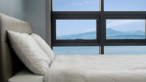 A bedroom with a bed, a pillow, and a window with a view of mountains and water. - Jigeum Yeosu Ocean View Pension (Yeosu)
