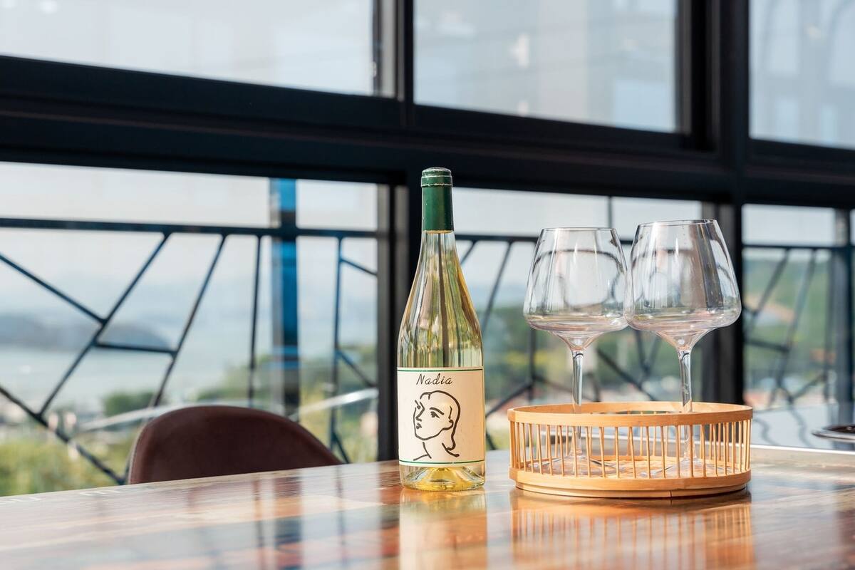 Een fles Nadja wijn en twee glazen op een houten dienblad, met uitzicht op zee door grote ramen.