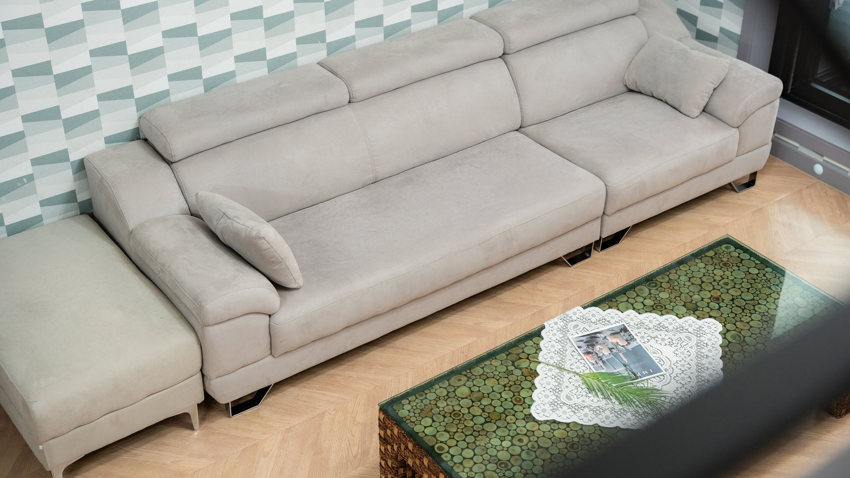 A beige sofa with a matching ottoman against a patterned wall, a small coffee table with a lace doily and a picture, and a herringbone wooden floor.