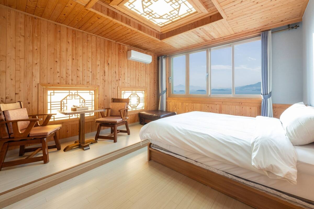 Een houten kamer met een bed, twee stoelen, een tafel en grote ramen met uitzicht.