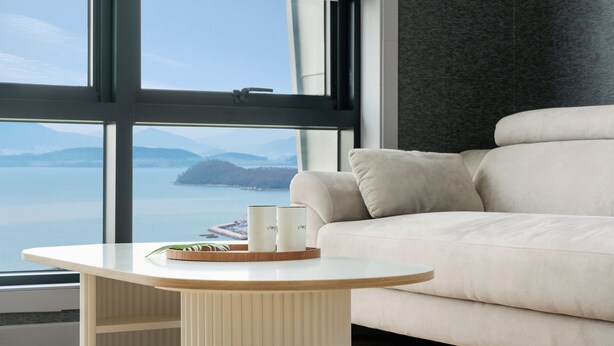 A modern living room with a large window, a beige sofa, and a round table with two white candles and a tray. - Jigeum Yeosu Ocean View Pension (Yeosu)