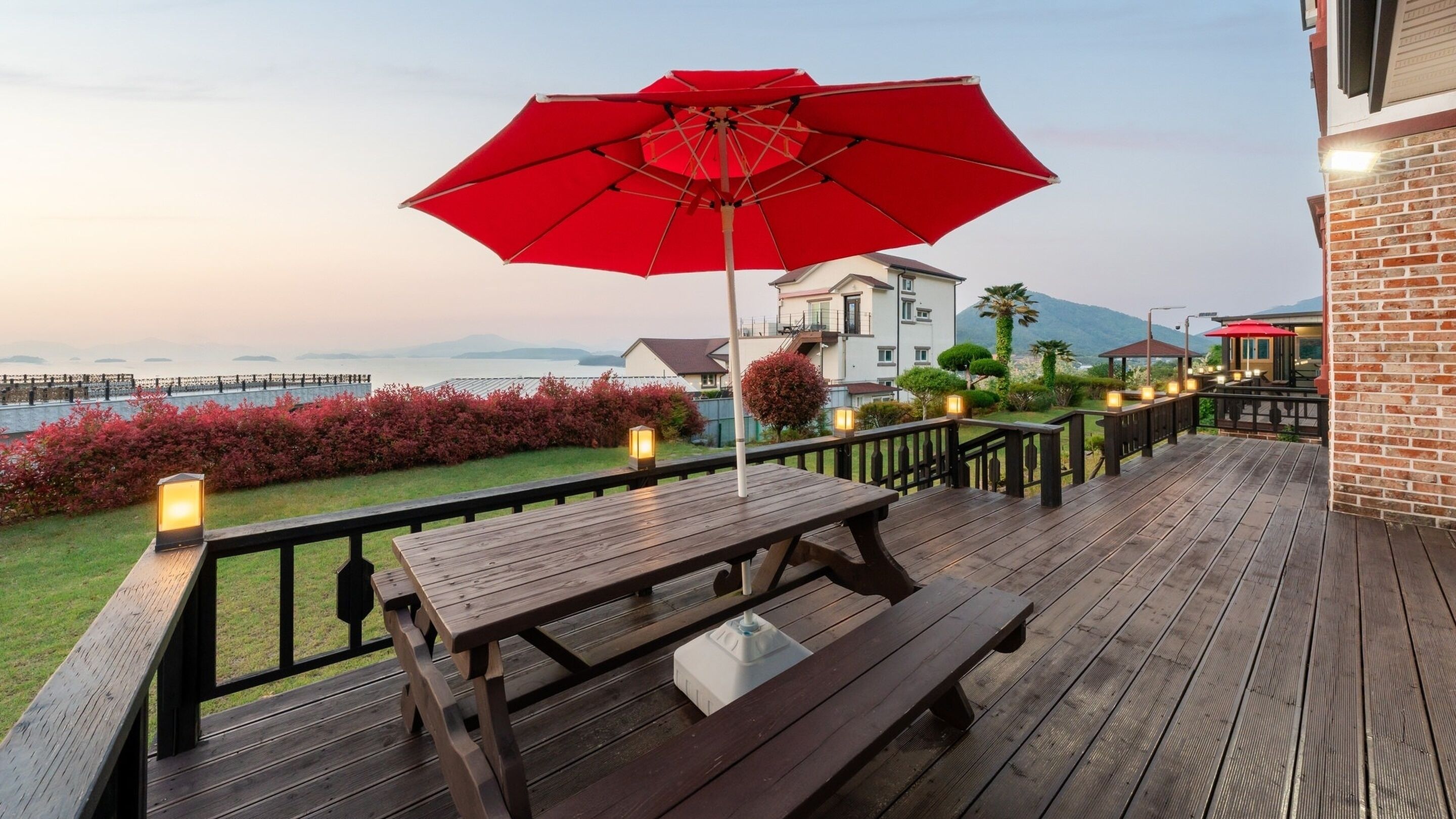 Une terrasse en bois avec des tables de pique-nique et un parasol rouge, donnant sur un jardin aménagé et un plan d’eau.