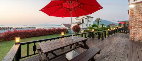 Une terrasse en bois avec des tables de pique-nique et un parasol rouge, donnant sur un jardin aménagé et un plan d’eau.