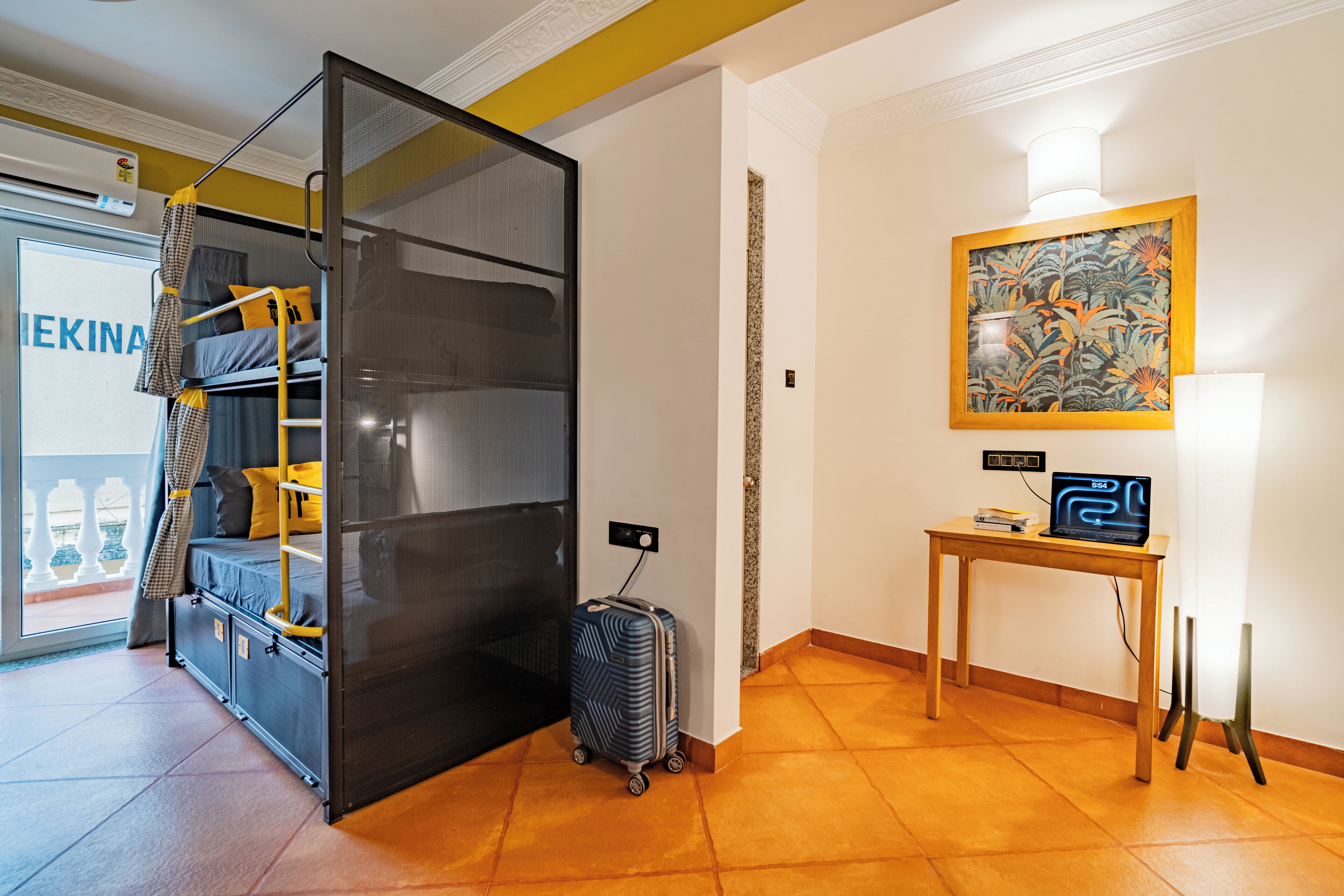 A room with a bunk bed, a suitcase, a desk with a laptop, and a painting on the wall.