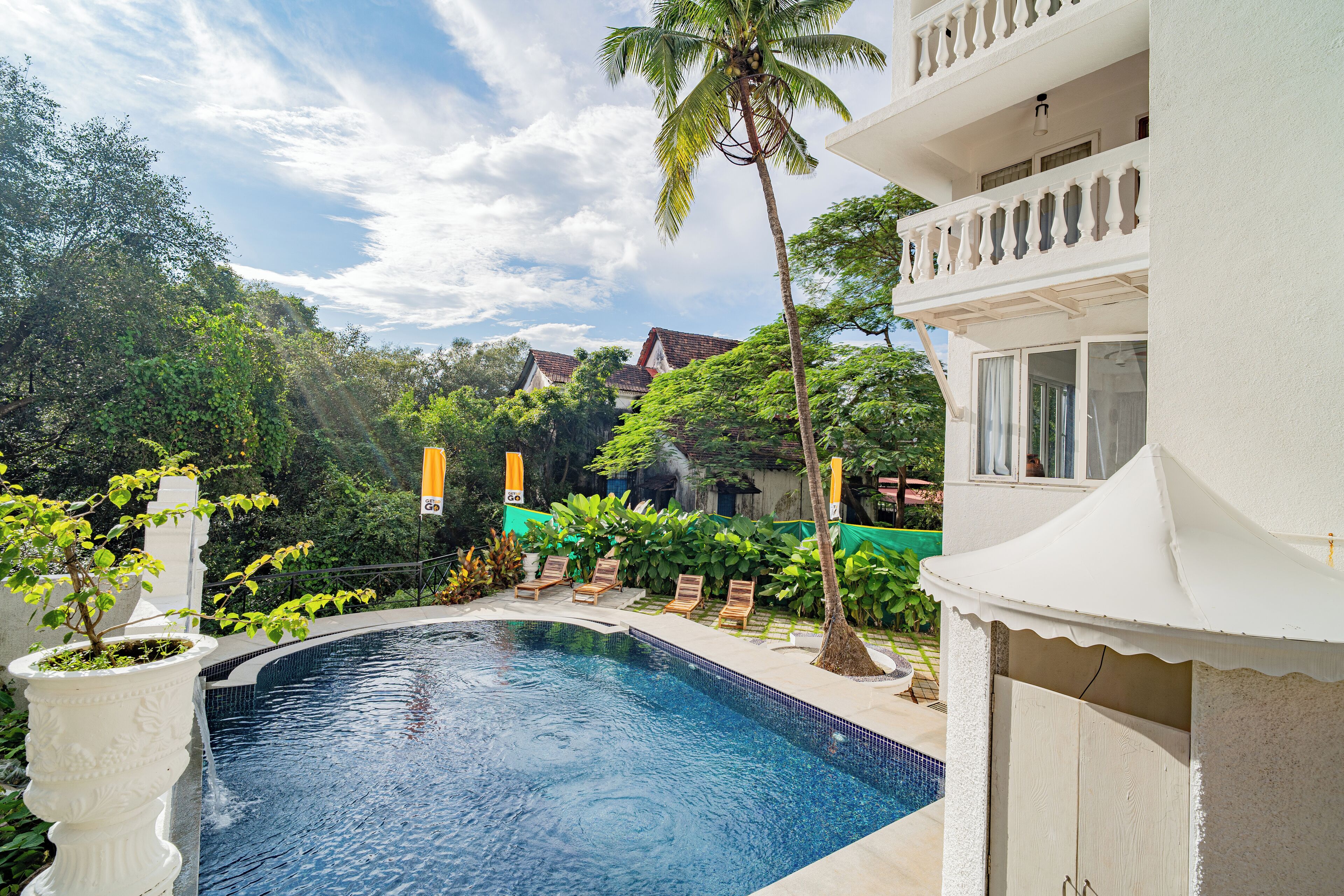A swimming pool with lounge chairs, surrounded by lush greenery and a white building with balconies.