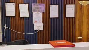 A reception desk with framed documents, a sign indicating advance check-out time, and a power outlet. - Hotel Sanjary International (Mumbai)