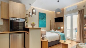 Ein kompaktes Hotelzimmer mit Küchenzeile, einem Bett mit blauer Decke, einem Fernseher und einem Balkon mit Blick.