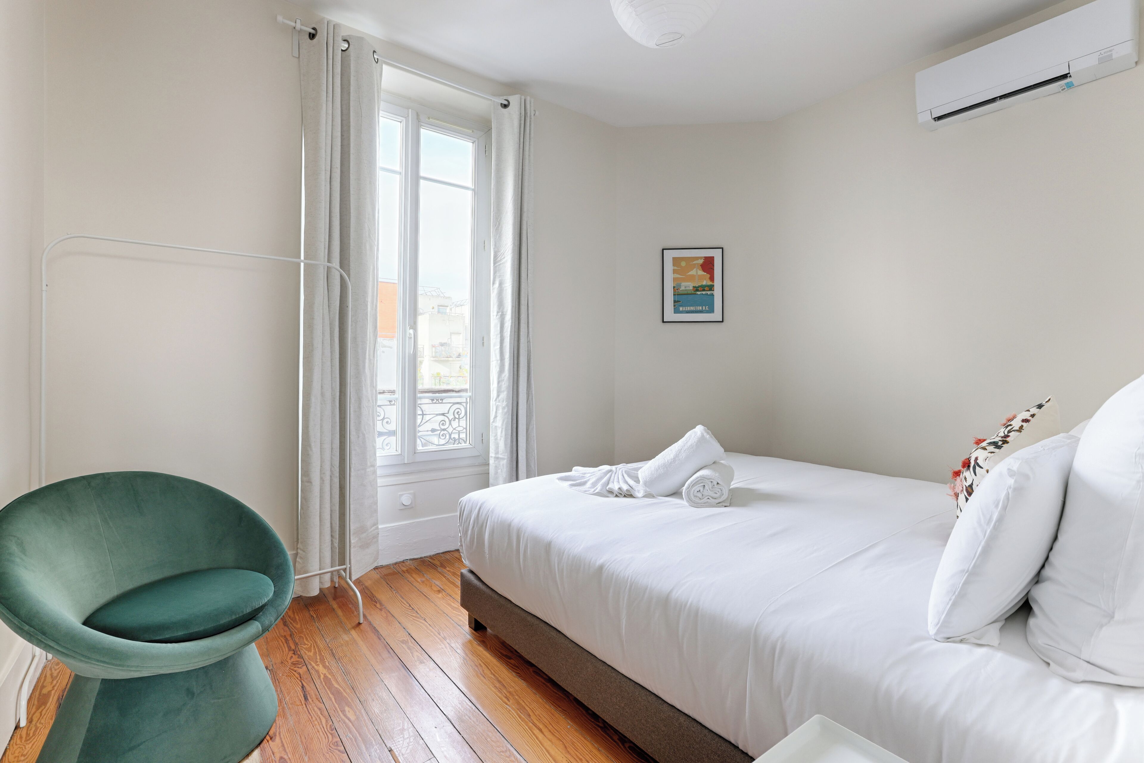 Une chambre à coucher avec un lit, un fauteuil vert, une fenêtre avec des rideaux et un appareil de climatisation.