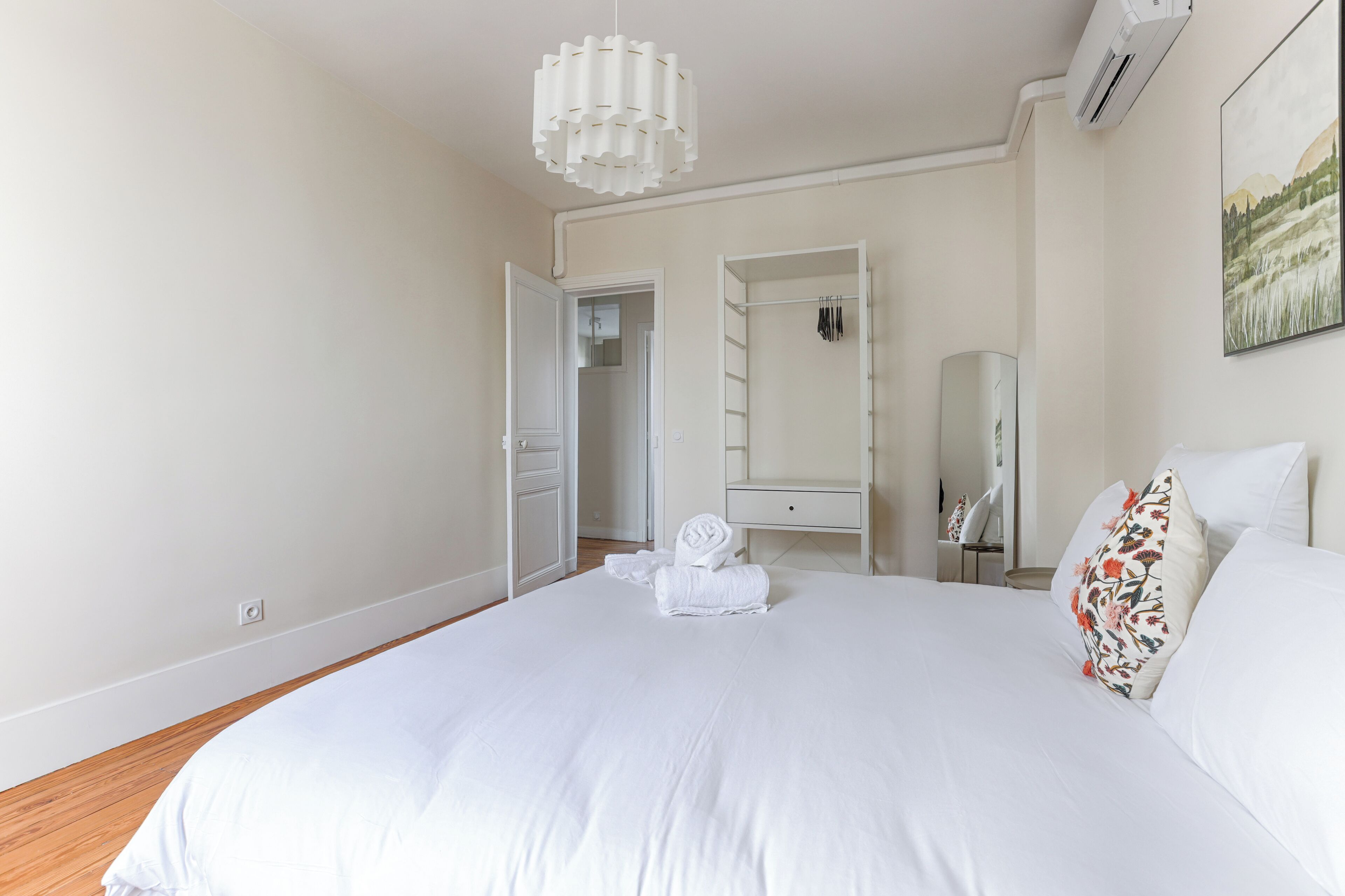 Een slaapkamer met een groot bed, wit beddengoed, een nachtkastje, een spiegel en een hanglamp.
