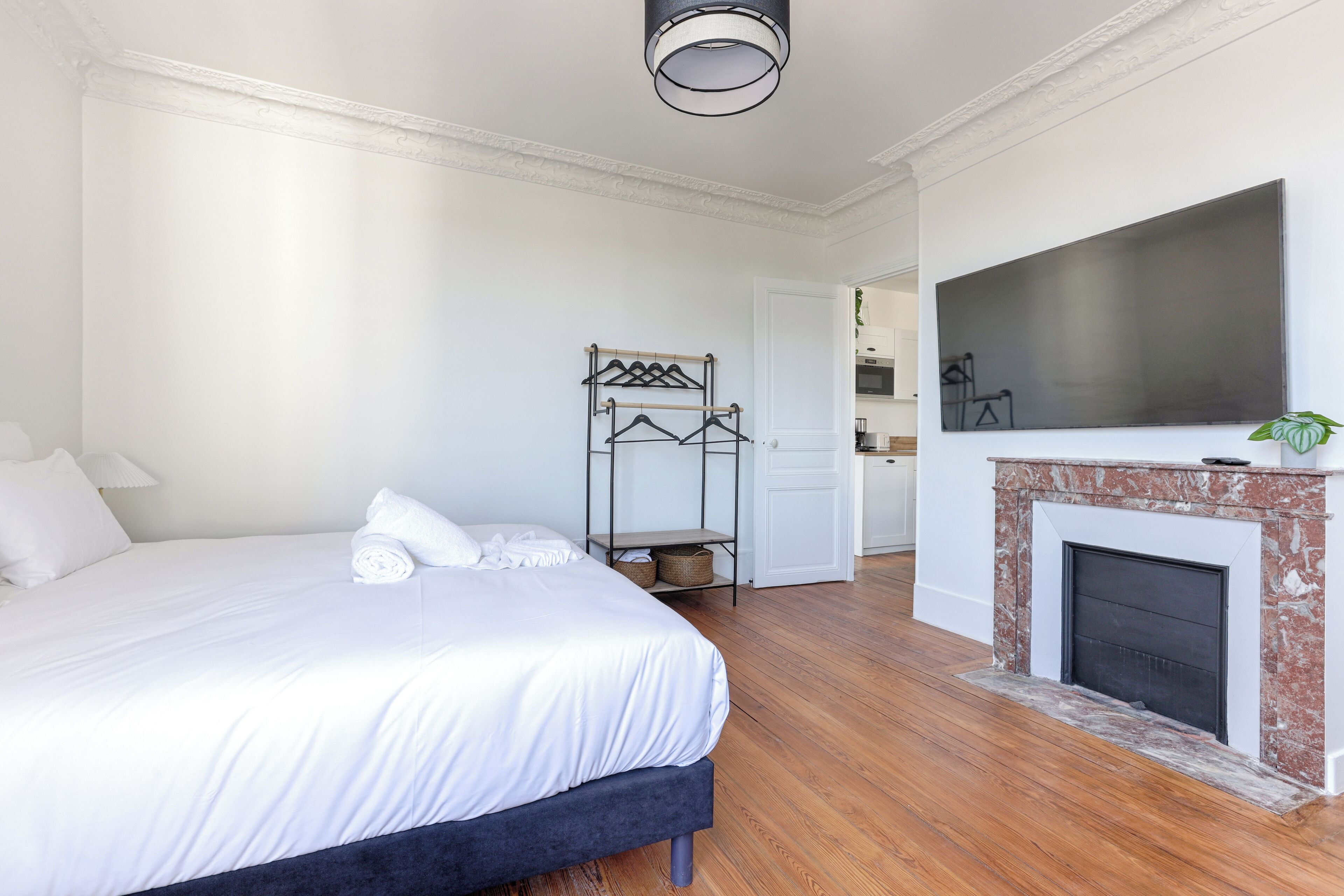 A bedroom with a bed, a hanging clothes rack, a fireplace, and a kitchen in the background.