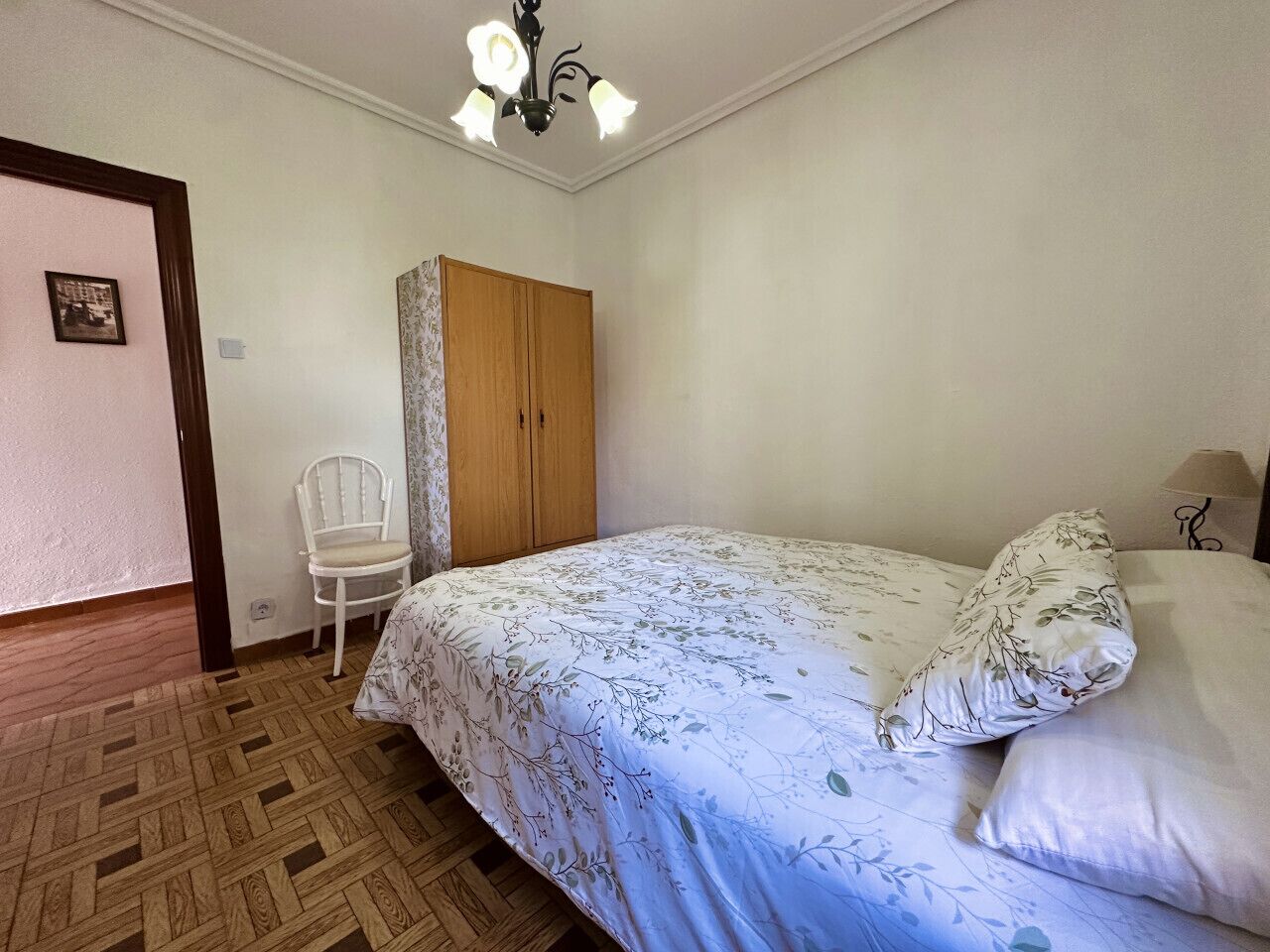 A bedroom with a bed, a wooden wardrobe, a white chair, and a chandelier.