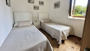 Ein Schlafzimmer mit zwei Betten, einem Nachttisch, einer Lampe und einem Fenster mit Blick auf Grünflächen.
