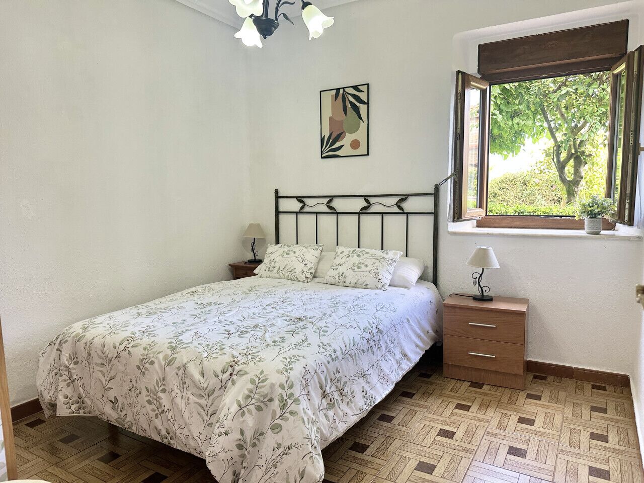 A bedroom with a bed, a nightstand, a dresser, a window with a view of trees, and a wall-mounted picture.