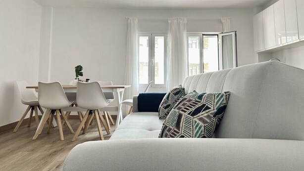 Een moderne woonkamer met een grijze bank, een eettafel met stoelen en een raam met gordijnen.