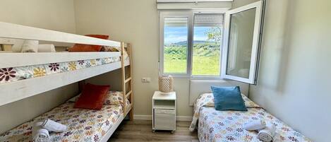 A small, well-lit room with two single beds, a window with a view, and a small table.