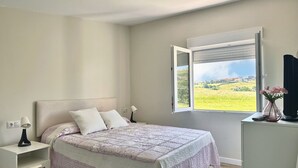 Ein Schlafzimmer mit Bett, Nachttisch, Lampe und einem Fenster mit Blick auf Grünflächen.