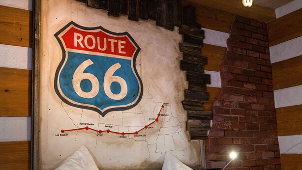Ett rum med en säng, kuddar, en skylt med texten ”Route 66”, en tegelbeklädd öppen spis och en hängande lampa.