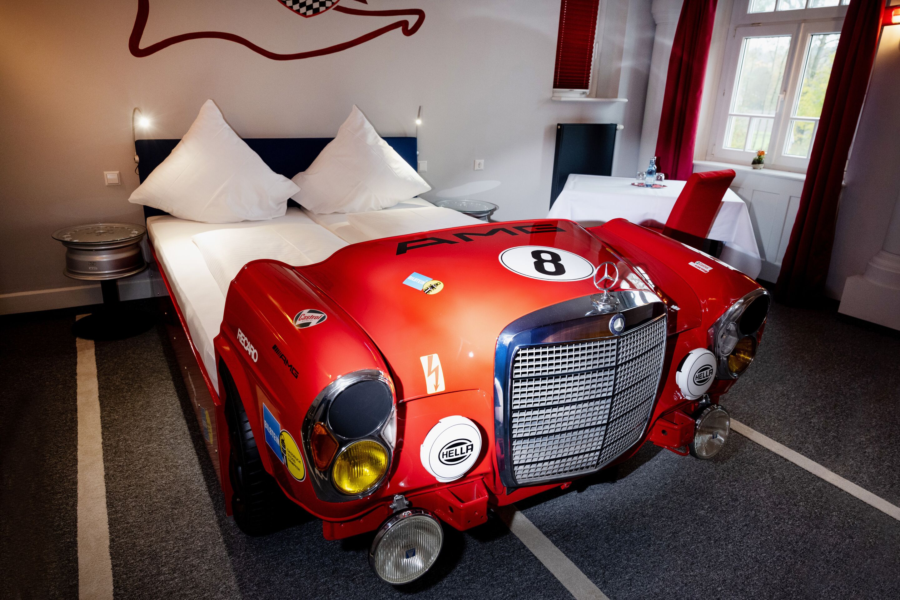 A hotel room with a bed, a red race car-shaped headboard, and a window with red curtains.