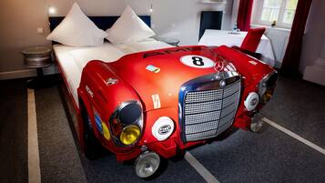 A hotel room with a bed, a red race car-shaped headboard, and a window with red curtains.