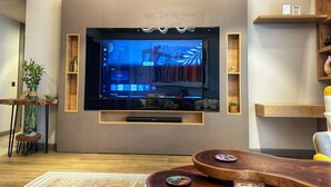 A modern living room with a large flat-screen TV mounted on a wooden wall, a small side table with a plant, and a round wooden coffee table with remote controls. - Charming 1-bedroom condo at The Diamond In City, Cantonments Accra. Luxe-Modern (Accra)