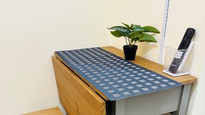 Un pequeño escritorio de madera con mantel azul estampado, una planta en maceta y un control remoto sobre un soporte.