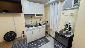 Una cocina compacta con armarios blancos, un fregadero, una estufa y un refrigerador pequeño. Hay un reloj en la pared y una planta en la encimera.