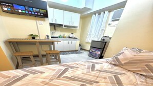 Habitación de hotel compacta con cama, pequeña cocina, escritorio y televisión montada en la pared.