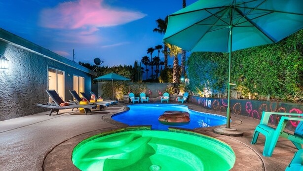 A swimming pool with lounge chairs and an umbrella, surrounded by greenery and a building. - Palm Springs Chic Retro Retreat w Pool/Spa/Games (Palm Springs)
