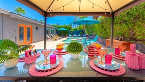 Un área de comedor al aire libre con piscina, sillones de descanso y una mesa dispuesta con vajilla rosa y bebidas.
