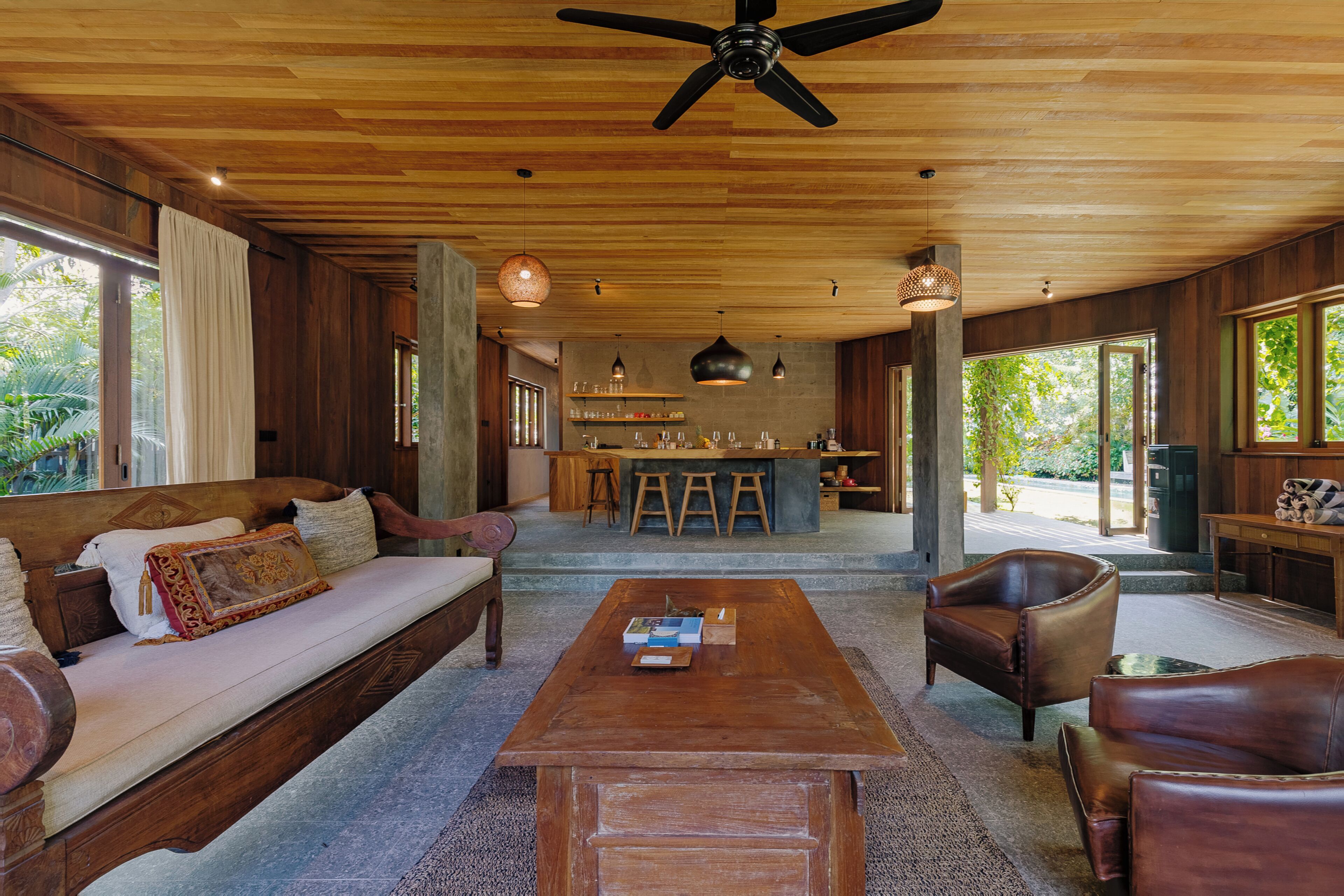 Amplia sala de estar con techo de madera, un sofá, una mesa de centro y sillas de cuero. Se ve una cocina al fondo con taburetes de bar.