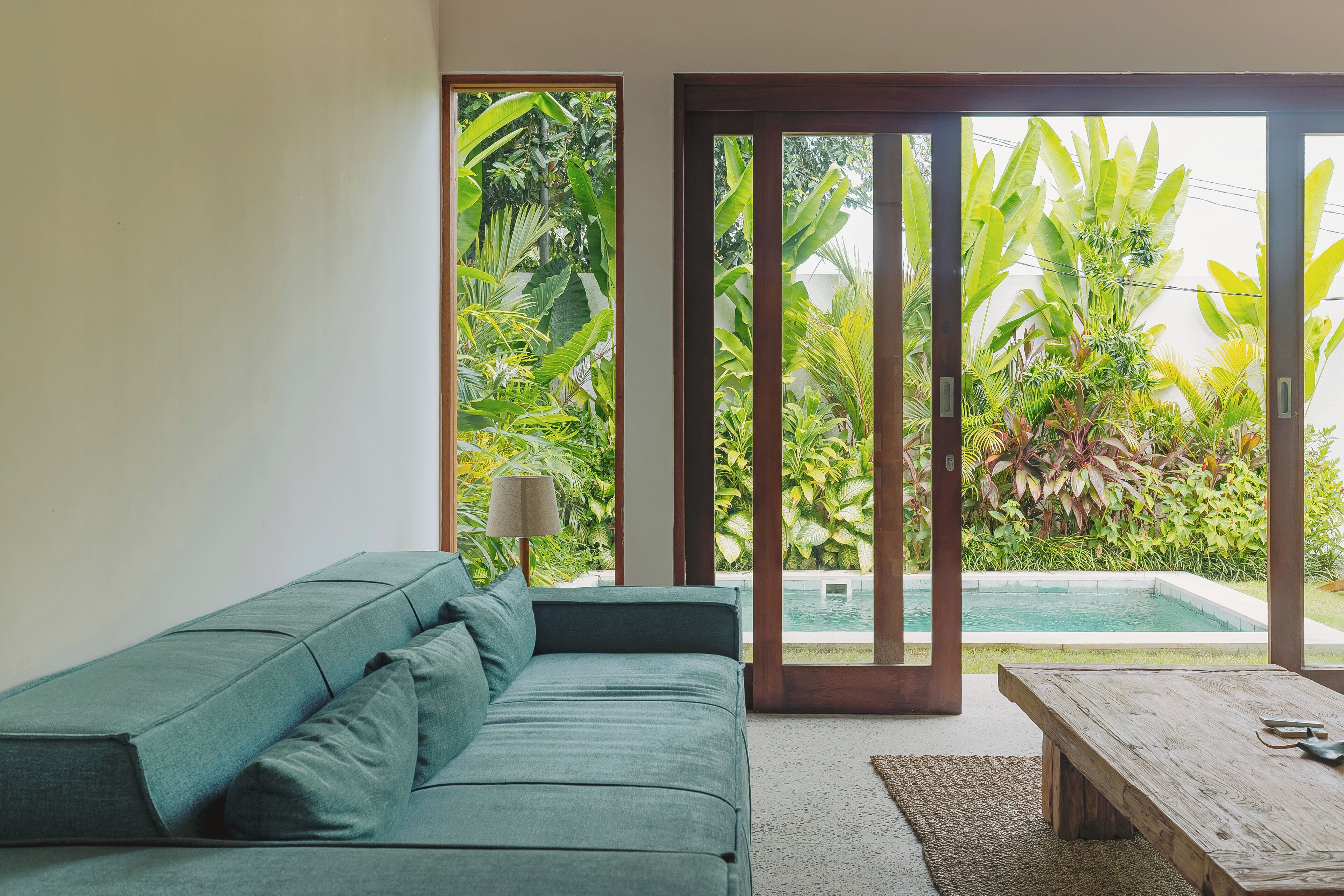 A modern living room with a teal sofa, a wooden coffee table, and a view of a swimming pool through large glass doors.