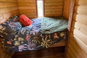 A small, wooden cabin room with a bed covered in a floral-patterned bedspread, a red pillow, and a green blanket.