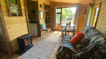 A cozy cabin interior with a sofa, a small table, a chair, a fireplace, and a window overlooking greenery.